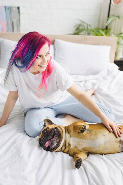 high angle view of girl with colorful hair petting cute french bulldog and looking away while sitting in bed