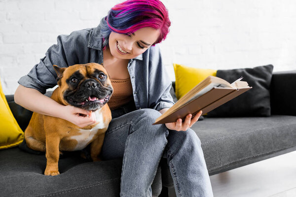 beautiful girl with colorful hair hugging bulldog, holding book, smiling and sitting on sofa