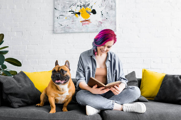 beautiful hipster girl sitting on sofa near small french bulldog and reading book