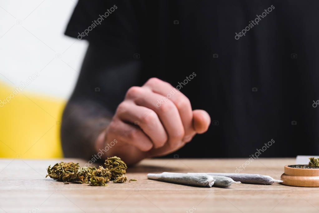 selective focus of weed, joints and herb grinder near man sitting at table