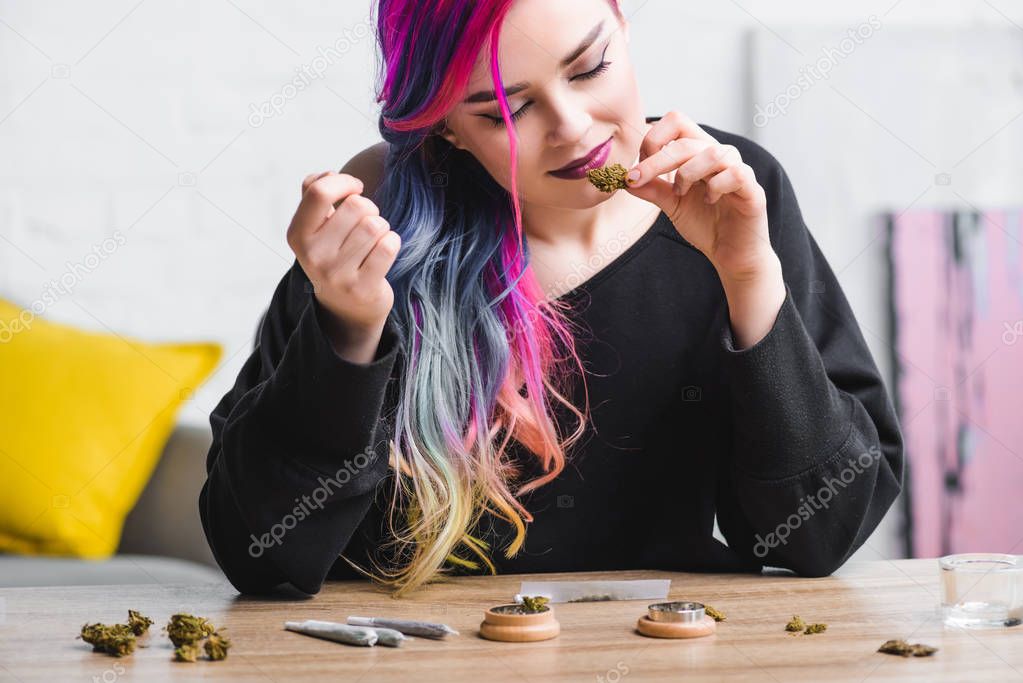 hipster girl with colorful hair sniffing medical marijuana and sitting behind table with joints and herb grinder 
