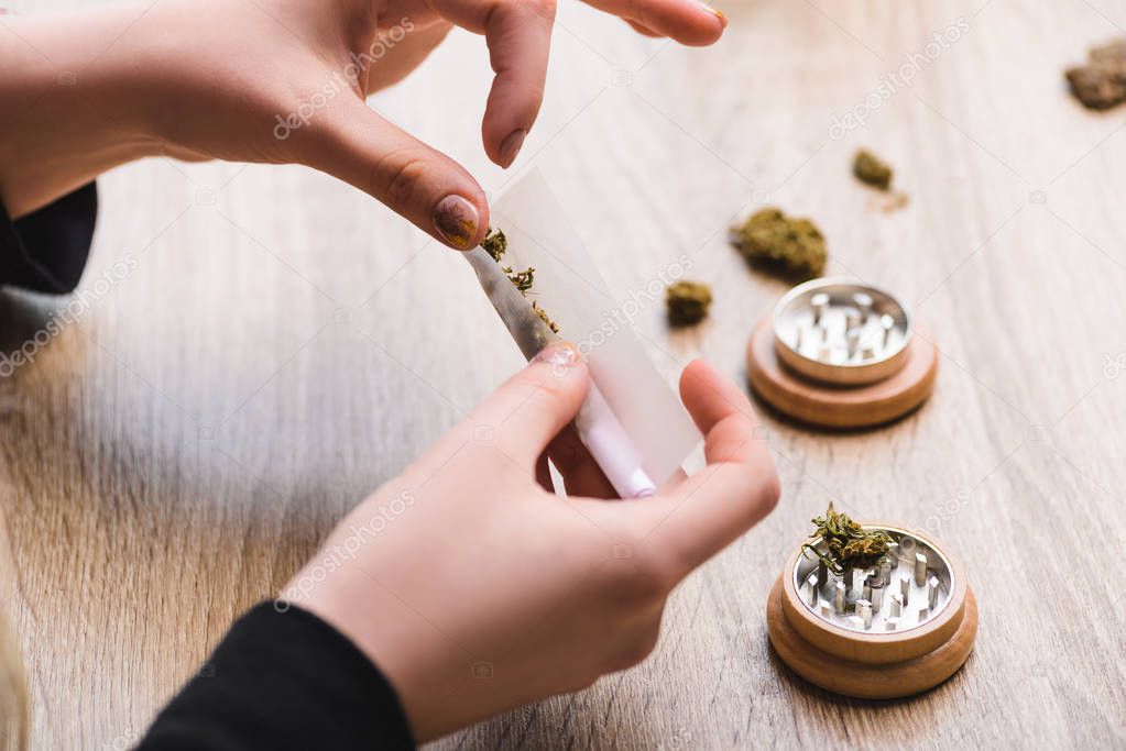 cropped view of woman making joint from weed and sitting at table with herb grinder 