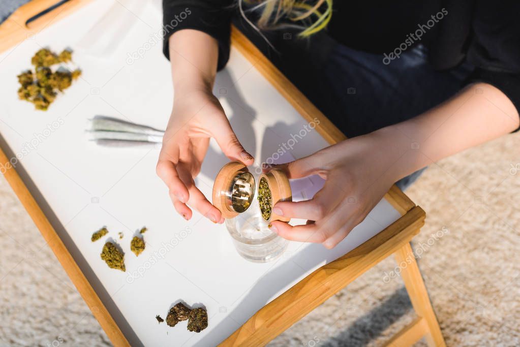 top view of girl putting weed from marijuana grinder in glass on wooden tray