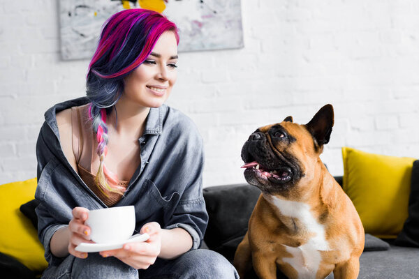 привлекательная девушка с красочными волосами держа чашку кофе и сидя на диване рядом с французским бульдогом
