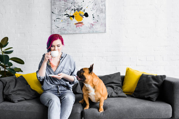 beautiful girl with colorful hair sitting on sofa and drinking coffee, while french bulldog looking at girl