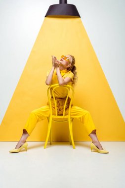 blonde woman in sunglasses sitting on chair and sending air kiss while looking at lamp on yellow and white clipart