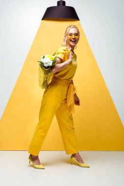 cheerful blonde woman holding string bag with flowers while posing on white and yellow  clipart
