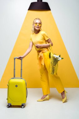 attractive blonde girl posing near luggage and holding reusable bag with flowers on white and yellow  clipart