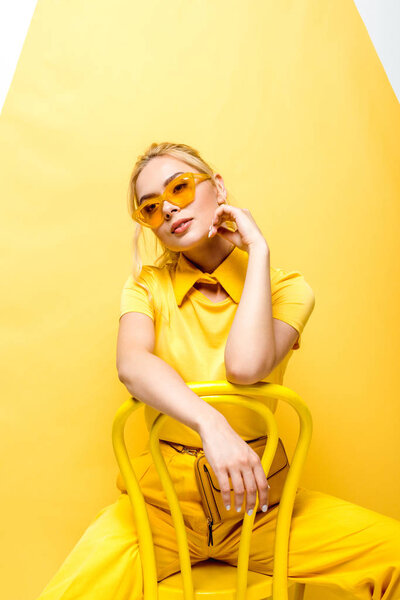 dreamy blonde woman in sunglasses sitting on chair on yellow