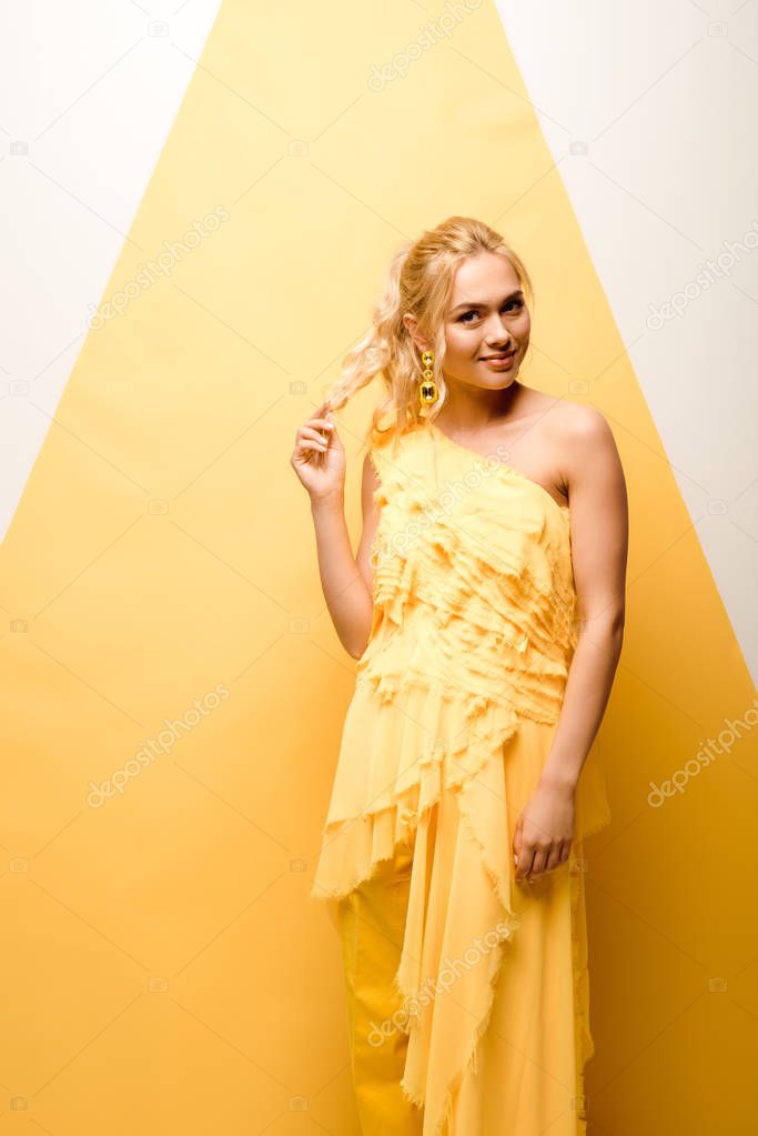 cheerful blonde girl touching hair while posing on white and yellow 