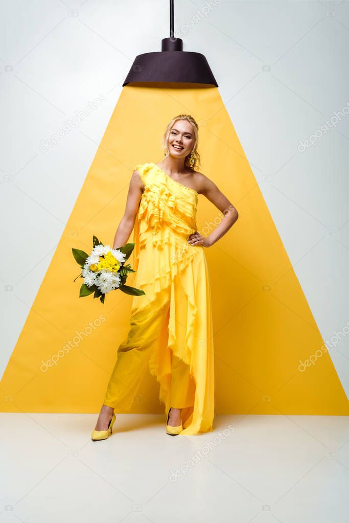 positive young woman holding bouquet of flowers and standing with hand on hip on white and yellow 