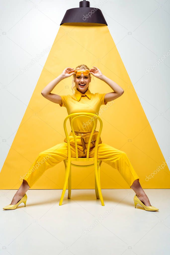 cheerful blonde woman touching sunglasses and sitting on chair on white and yellow 