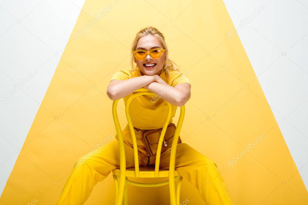 cheerful blonde woman in sunglasses sitting on chair on white and yellow 