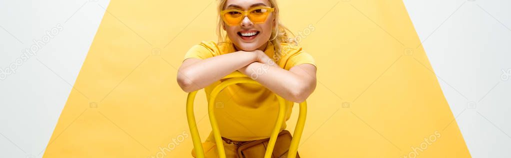 panoramic shot of happy blonde woman in sunglasses sitting on chair on yellow and white