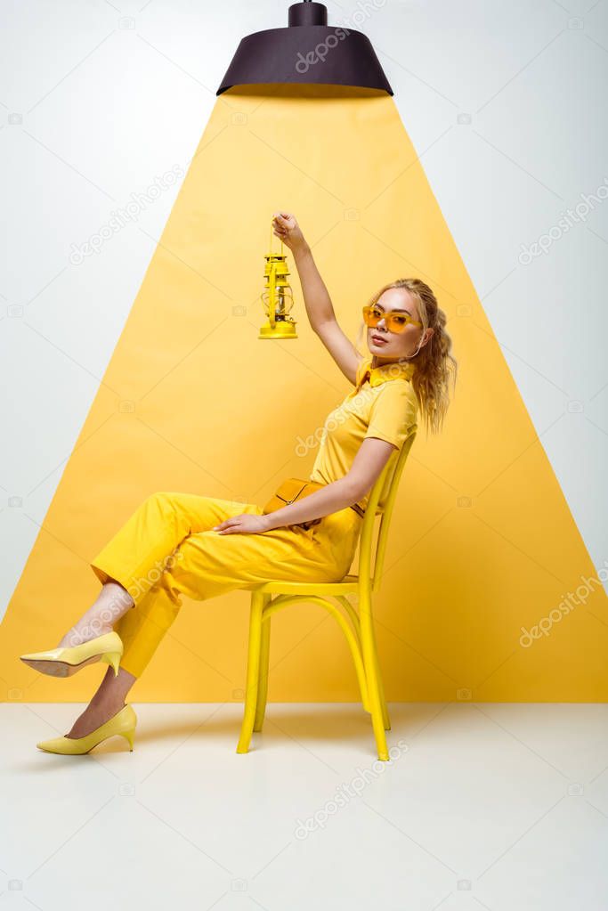 attractive blonde woman in sunglasses sitting on chair and holding vintage lamp on white and yellow 