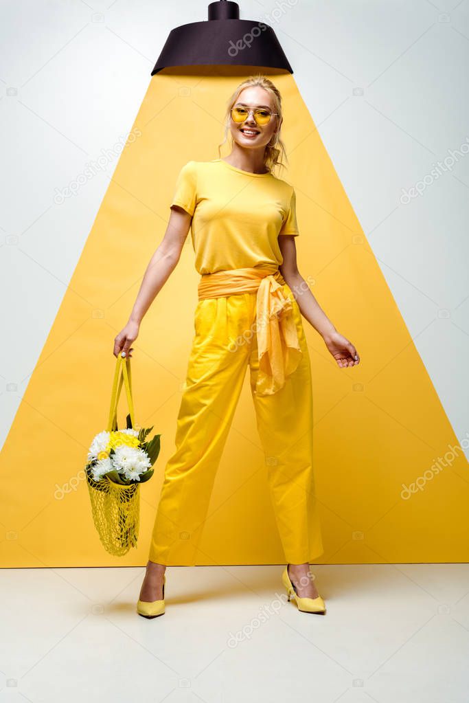 happy blonde woman holding string bag with flowers while posing on white and yellow 
