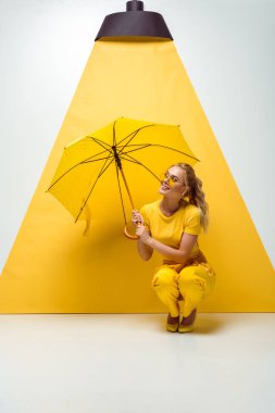 happy blonde girl holding umbrella while sitting on white and yellow  clipart