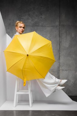 güzel genç kadın sandalyede oturan ve beyaz ve gri üzerinde sarı şemsiye tutan 