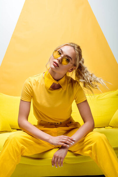attractive blonde woman in sunglasses sitting on sofa and looking at camera on white and yellow 