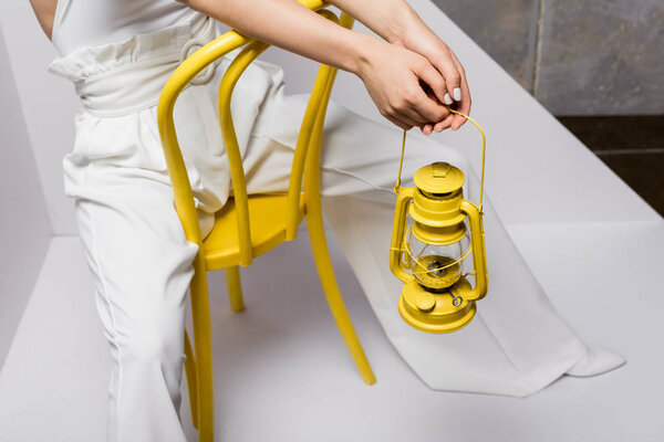 cropped view of young woman sitting on yellow chair and holding retro lamp on white and grey 