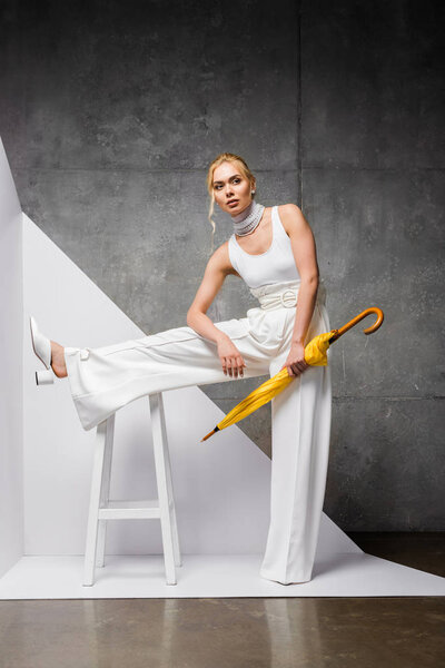 stylish pretty woman posing near chair with yellow umbrella on white and grey 