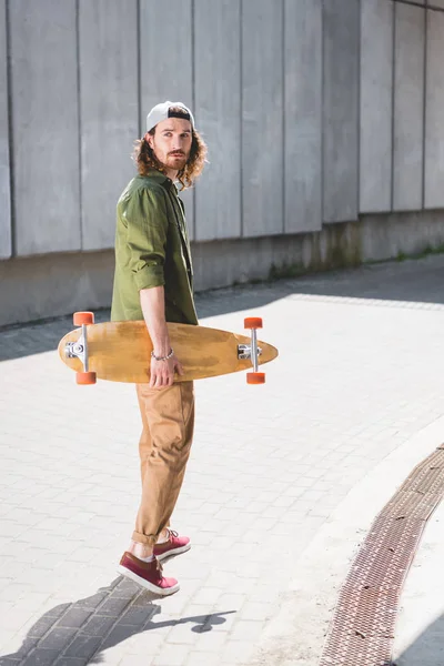 スケートボードを手にして集中壁の近くに立ってカジュアルな服装のハンサムな男 目をそらす — ストック写真
