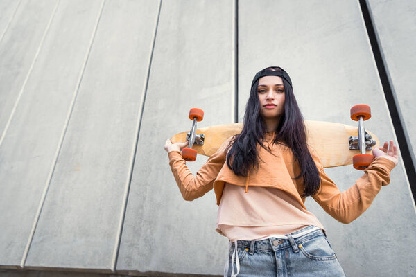 low angle view of brunette woman in casual wear standing near concentrate wall, holding skateboard behind back, looking at camera
