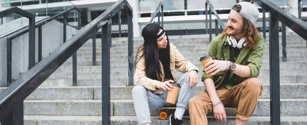 elinde kağıt bardak ile kaykay üzerinde oturan mutlu çift düşük açı görünümü, birbirlerine bakarak