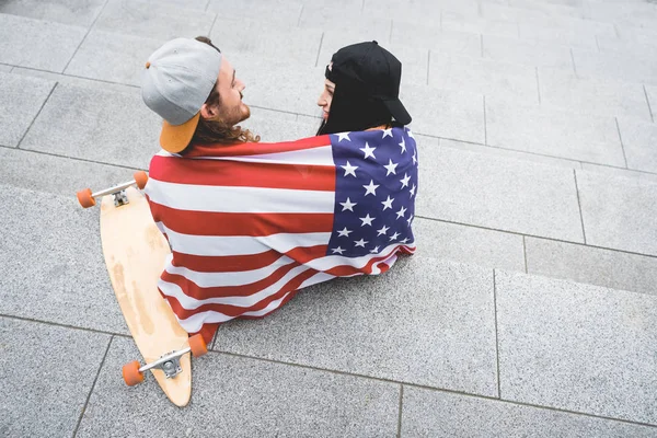 スケートボードの近くの階段に座って お互いを見て 肩にアメリカの旗を持つカップルの高い角度のビュー — ストック写真