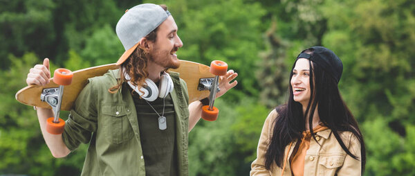 panoramic view of smiling man with skateboards looking at woman, standing on green trees background