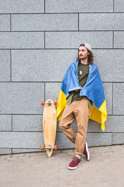 calm man in casual wear and ukrainian flag standing near wall with skateboard