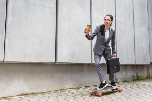 快乐的女商人在正式穿着骑在滑板上 手里拿着纸杯和公文包 — 图库照片
