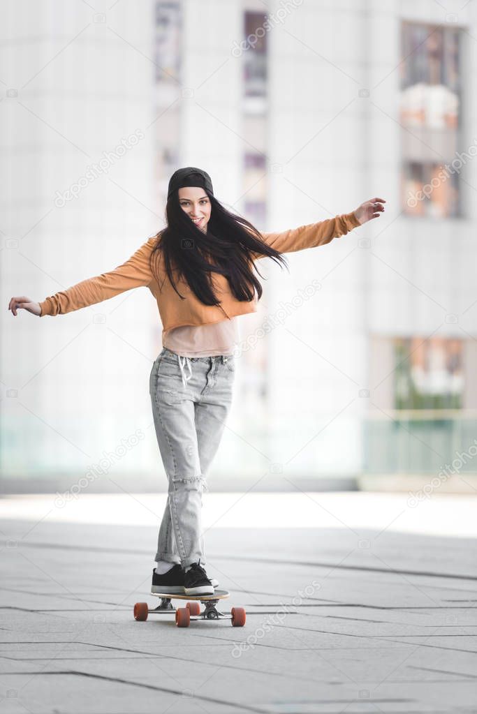 brunette woman with outstretched hands looking at camera, riding on skateboard in city