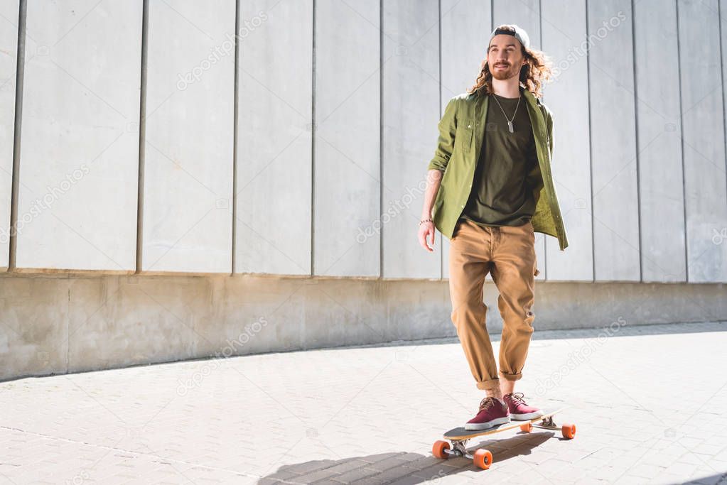 low angle view of handsome man looking away, riding on skateboard near concrete wall