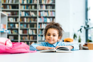 mutlu afrikalı amerikalı çocuk oturan ve kitap ve pembe sırt çantası yakınında kamera bakıyor 