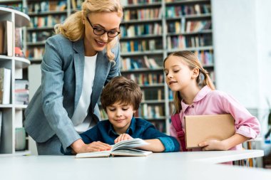 cheerful teacher in glasses standing near cute kids reading book in library  clipart