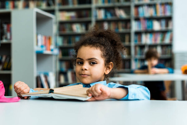 selective focus of happy african american kid sitting and looking at camera near book on table 