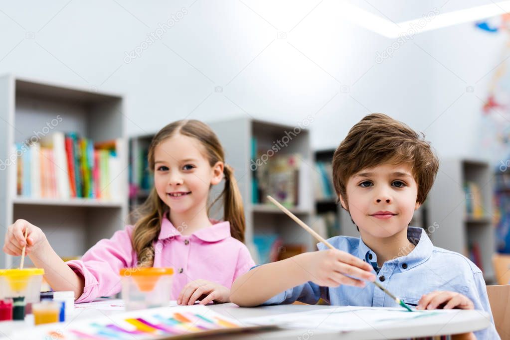 selective focus of cheerful kids looking at camera and holding paintbrushes 