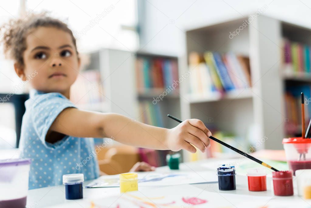 selective focus of african american child holding paintbrush near gouache jars