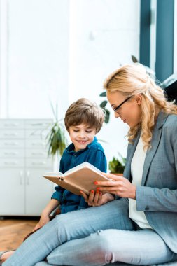 woman in glasses sitting on floor and reading book with cute kid  clipart