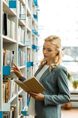 beautiful blonde woman reading book while standing in library  clipart