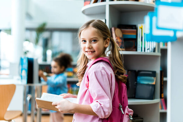 cheerful kid with pink backpack standing and holding book in library 