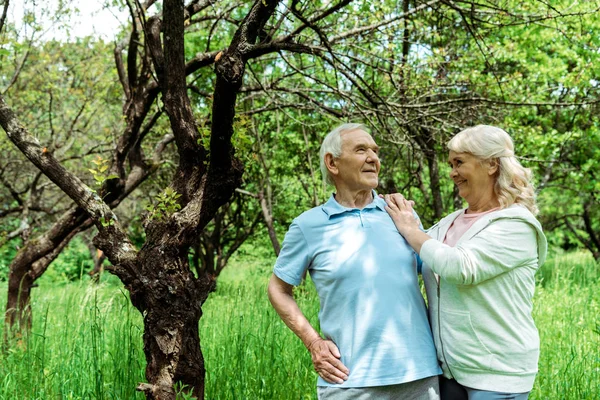 妻と緑の木の近くに腰に手を置いて立っている陽気な先輩男性 — ストック写真