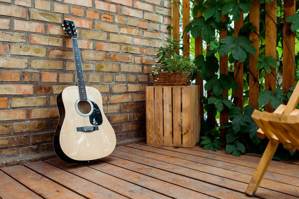 selective focus of acoustic guitar near brick wall and wooden fence with green leaves 