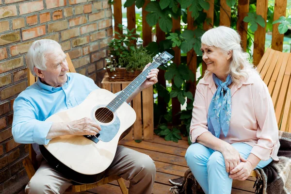 happy retired woman looking at husband playing acoustic guitar