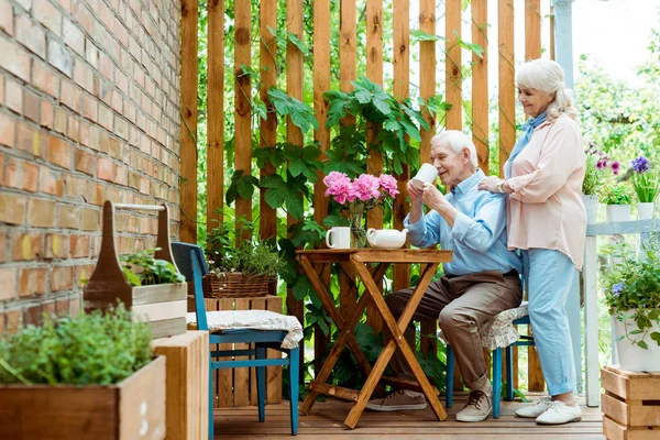 Selective focus of cheerful senior wife standing near husband drinking tea — Stock Image Selective Focus Cheerful Senior Wife Standing Husband Drinking Tea Stock Image