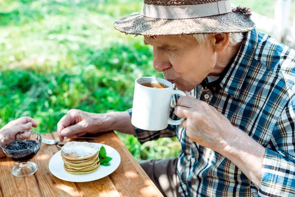 甘くておいしいパンケーキの近くでお茶を飲む麦わら帽子の先輩男性 — ストック写真
