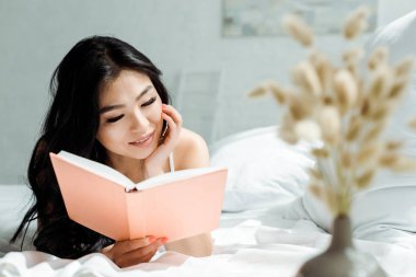 selective focus of positive asian girl reading book while lying on bed at home  clipart