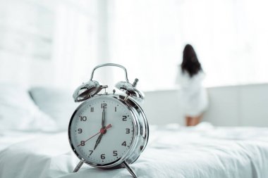 selective focus of alarm clock near brunette woman standing at home  clipart