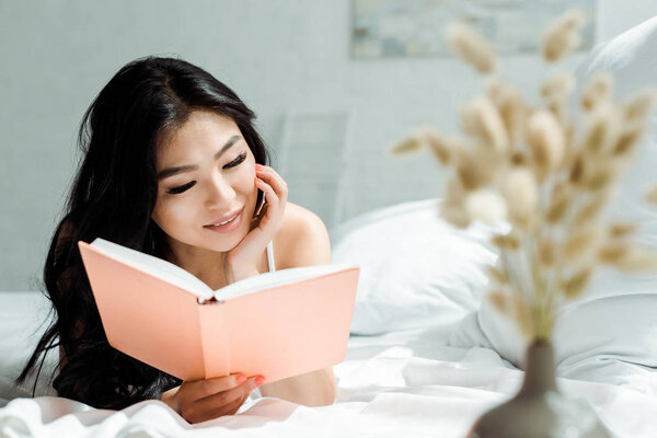 selective focus of positive asian girl reading book while lying on bed at home 